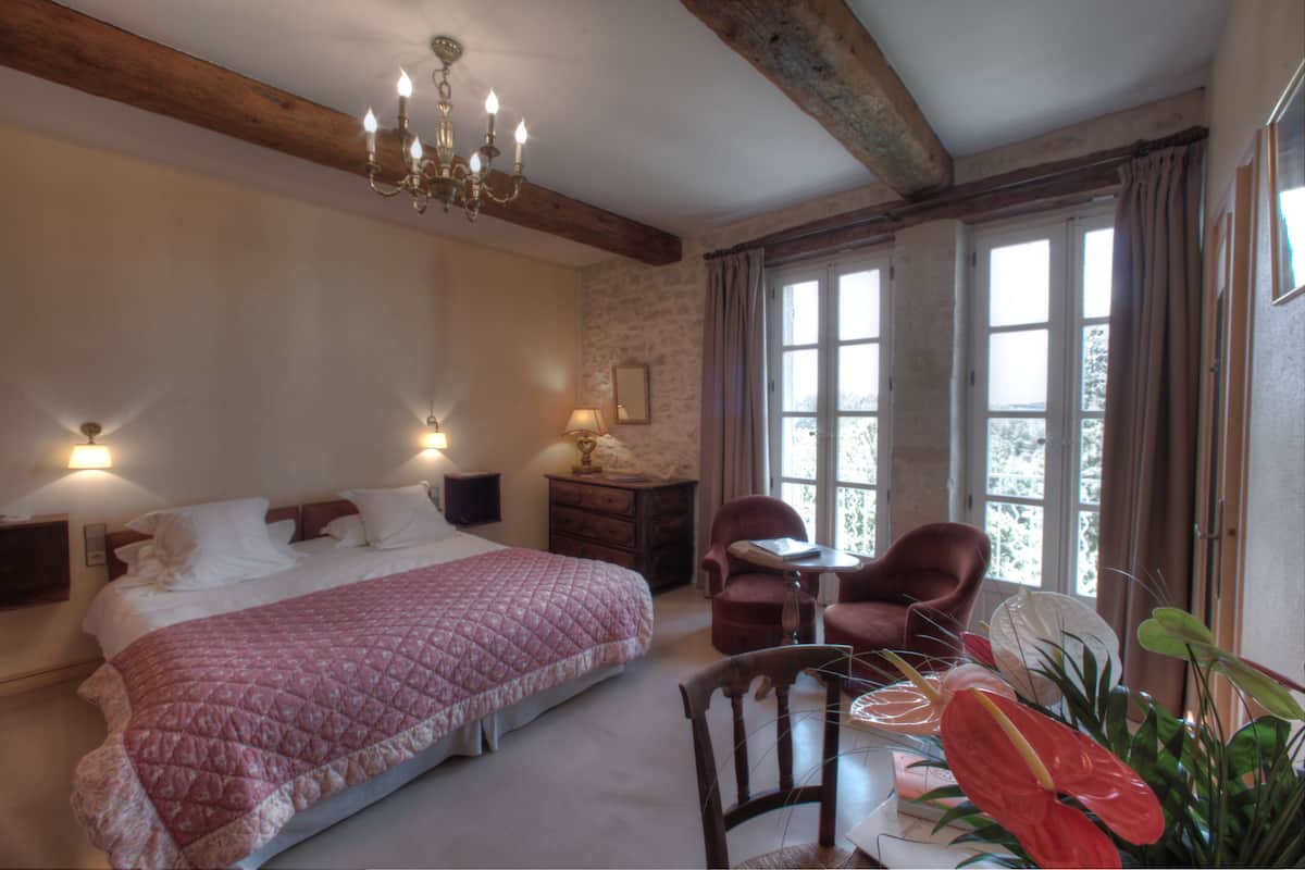 A bedroom with a large bed, a wooden ceiling, a chandelier, a desk with a chair, and a window with curtains.