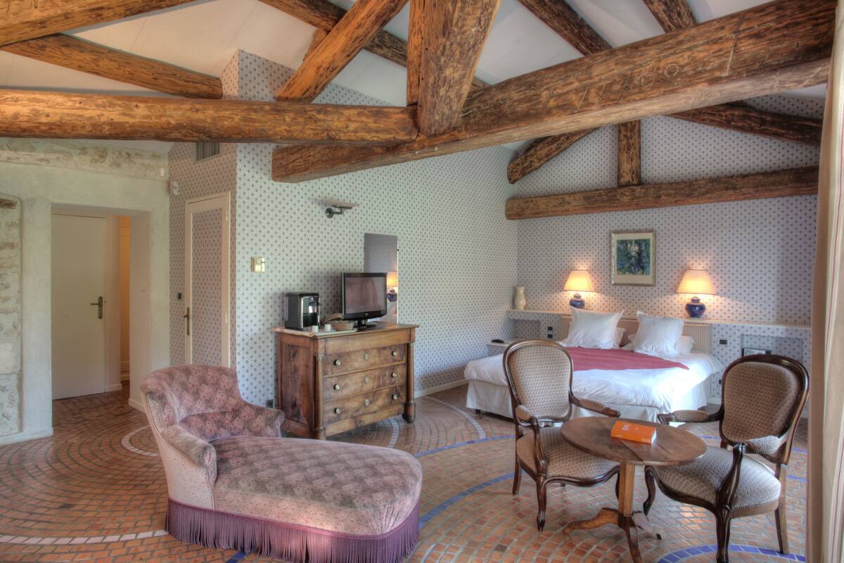 A hotel room with a wooden ceiling, a bed with white linens, two bedside tables with lamps, a wooden dresser, a television, and a patterned armchair.