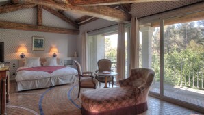 A bedroom with a large bed, two chairs, a table, and a balcony with a view of trees.