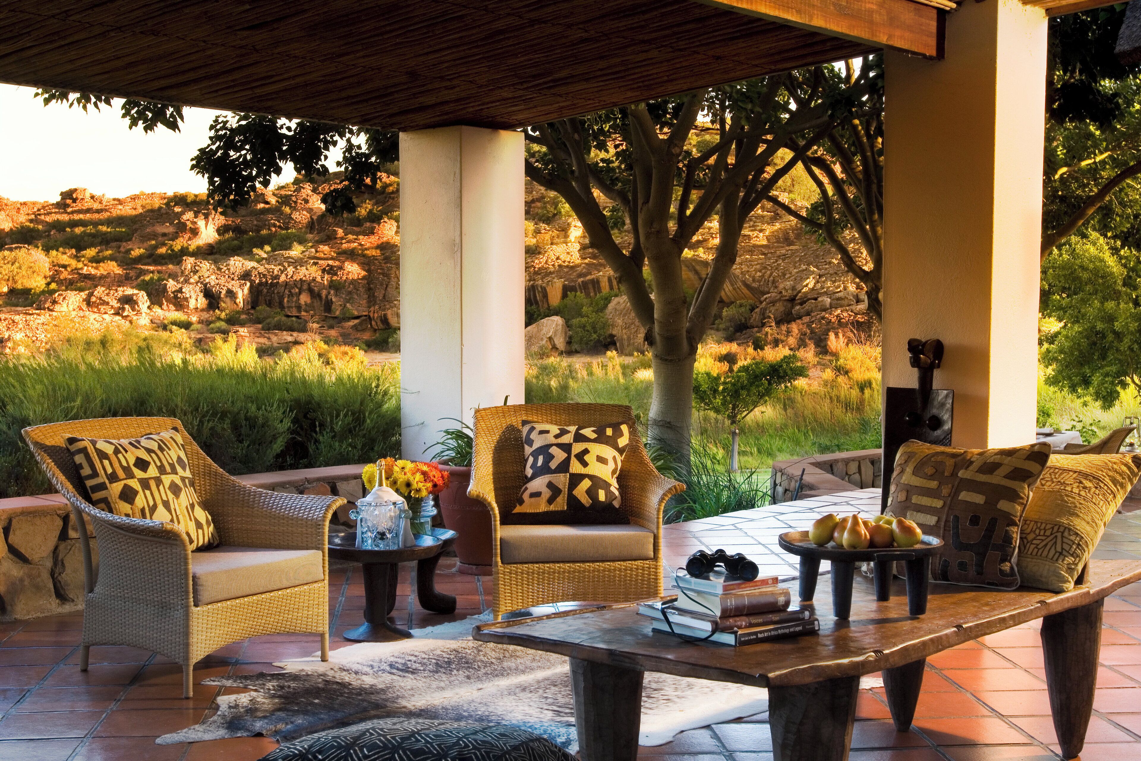 A covered outdoor seating area with wicker furniture, a coffee table with books and pastries, and a view of a natural landscape.