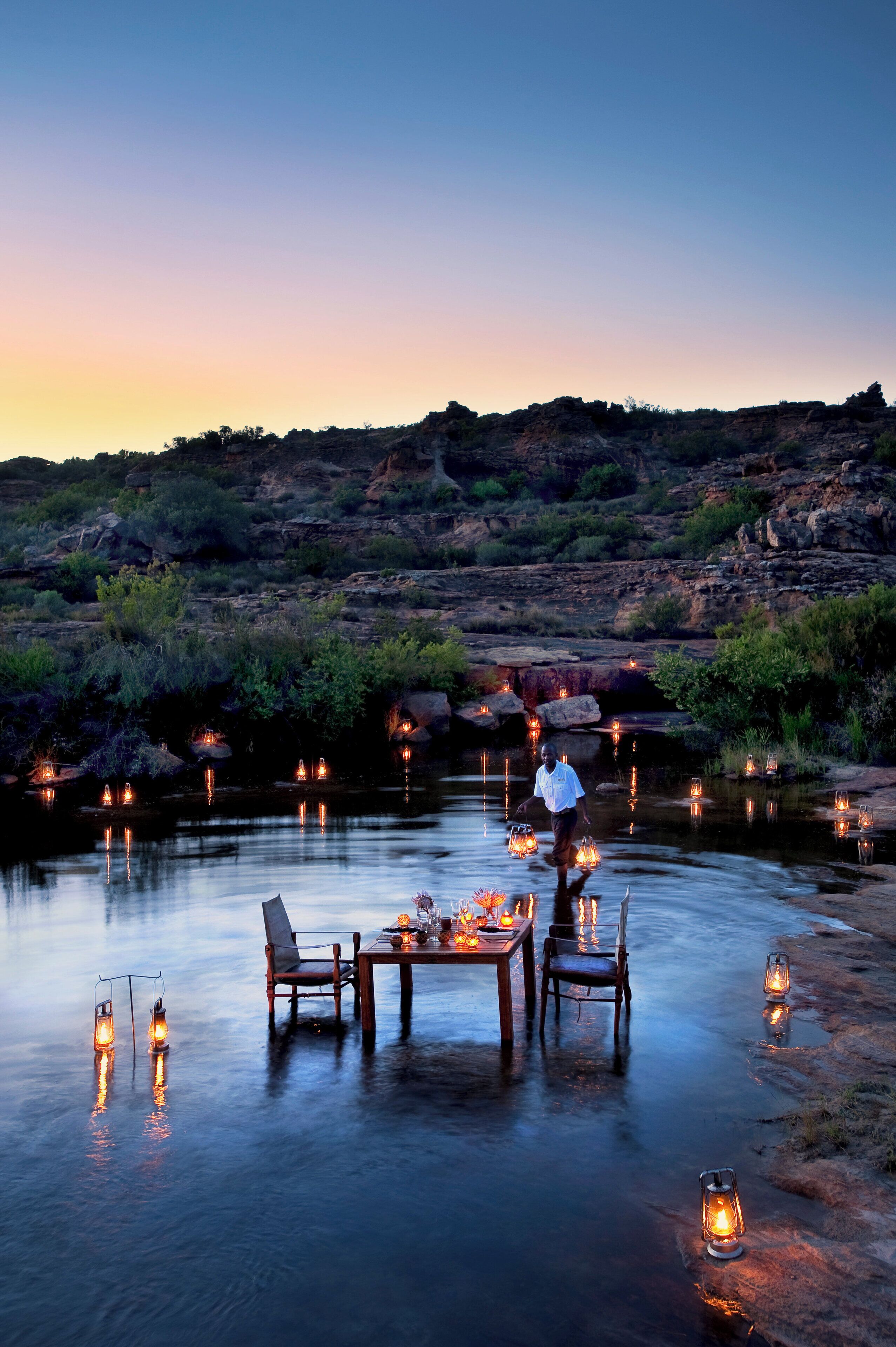 A serene outdoor dining area with a table and chairs set up on a rocky shore, surrounded by lanterns.
