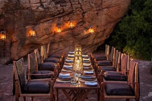 Una larga mesa de comedor preparada para una comida, con sillas a ambos lados, ubicada al aire libre frente a un fondo rocoso.