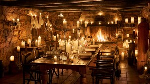 Une salle à manger faiblement éclairée, avec une longue table en bois dressée pour le repas, entourée de murs en pierre et illuminée par des bougies.