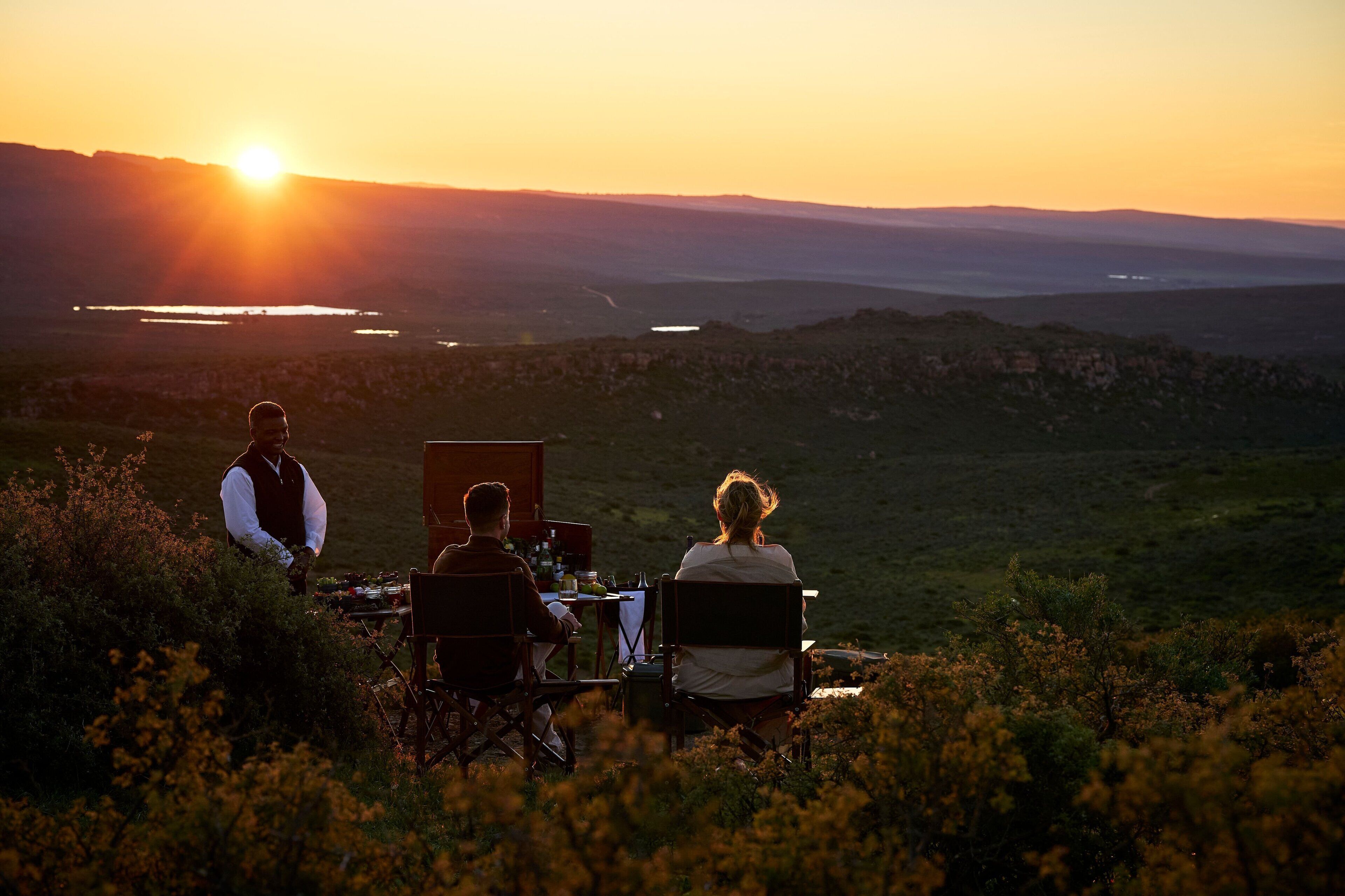 A couple dining outdoors with a scenic view of the sunset and hills in the background.