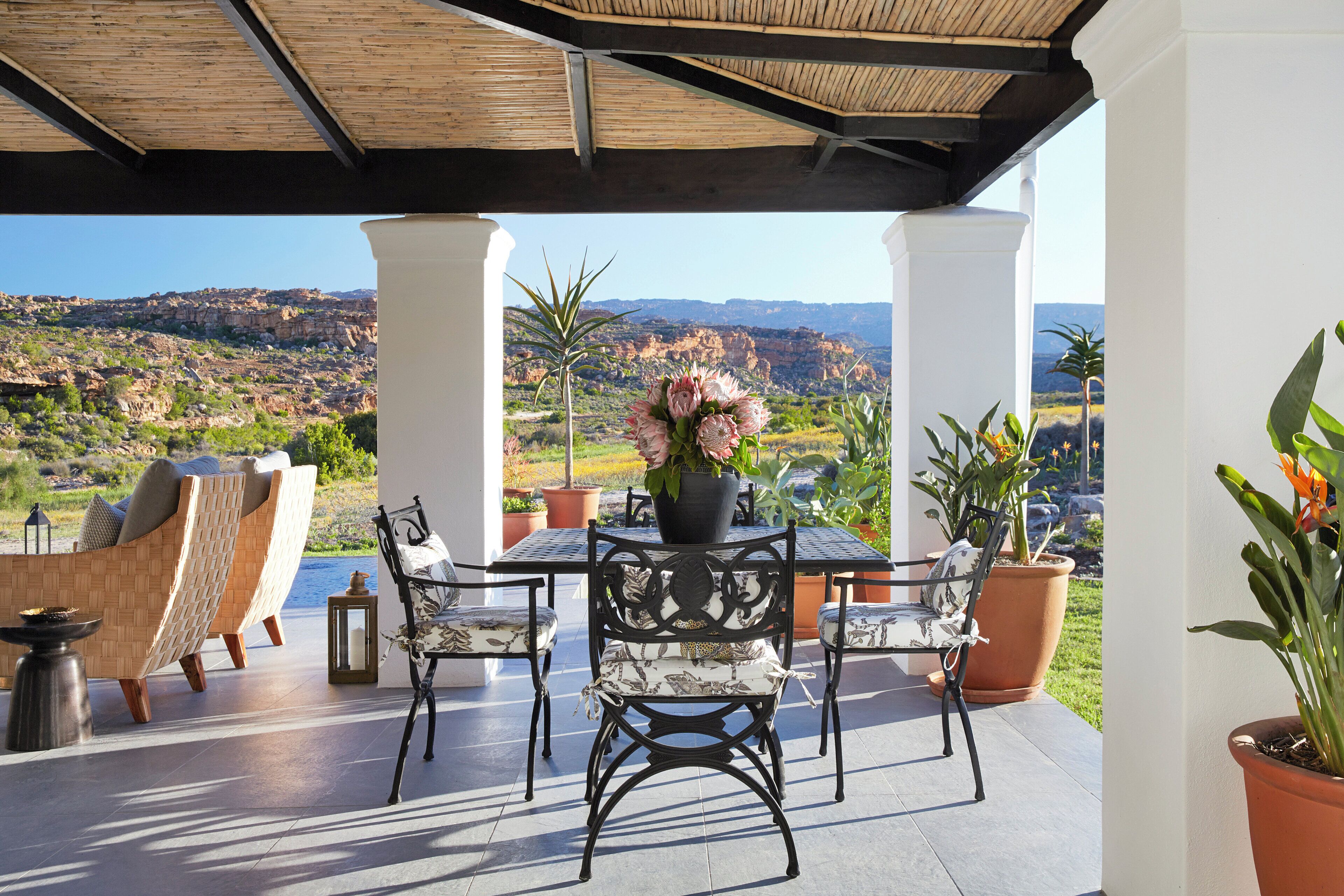 A patio with a table and chairs, surrounded by potted plants and a view of a mountainous landscape.