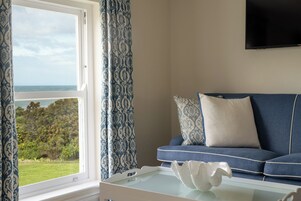 Una habitación con vista al mar a través de una ventana, un sofá azul con cojines estampados y un objeto decorativo blanco sobre la mesa.