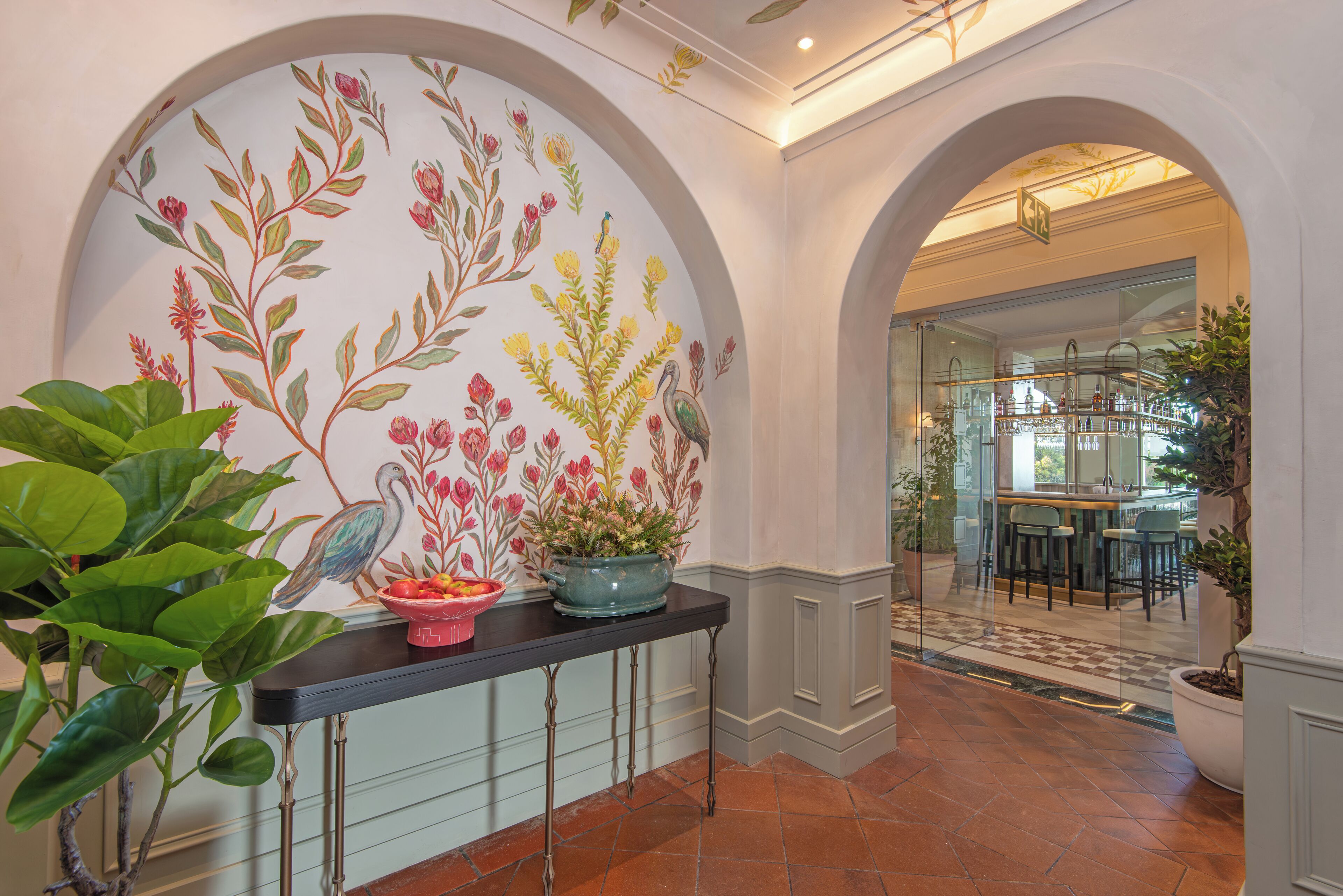 Un pasillo con un mural floral, una consola negra con plantas en macetas y un arco que conduce a un comedor.