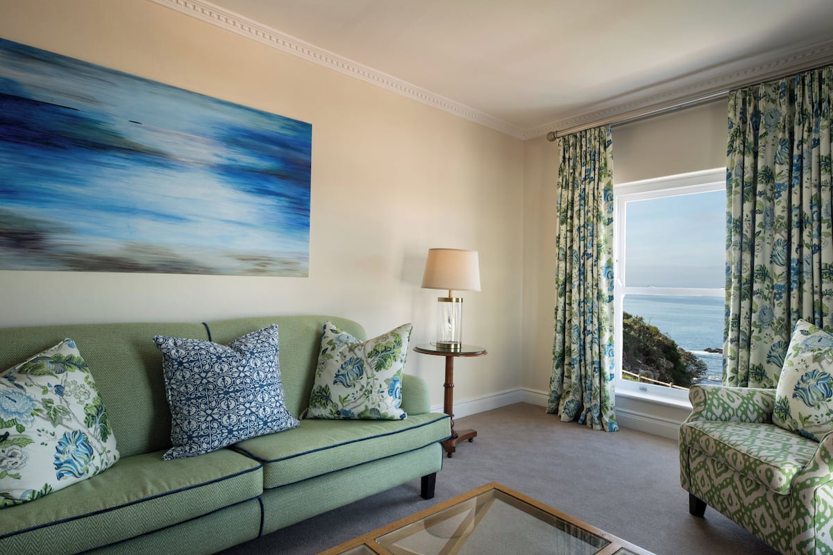A living room with a green sofa, patterned cushions, a side table with a lamp, and a view of the sea through curtains.