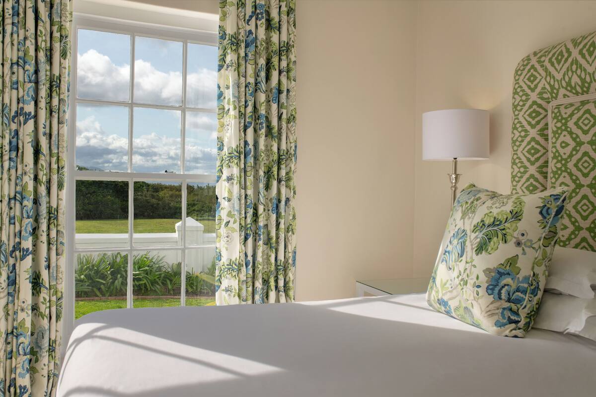 A bedroom with a large window, floral curtains, a bed with patterned pillows, and a bedside table with a lamp.