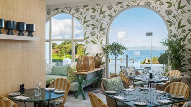 A dining area with tables and chairs, a view of the sea, and a balcony with plants.