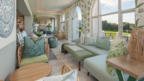 A spacious room with a large window, a sofa, a round table, and a dining area in the background.