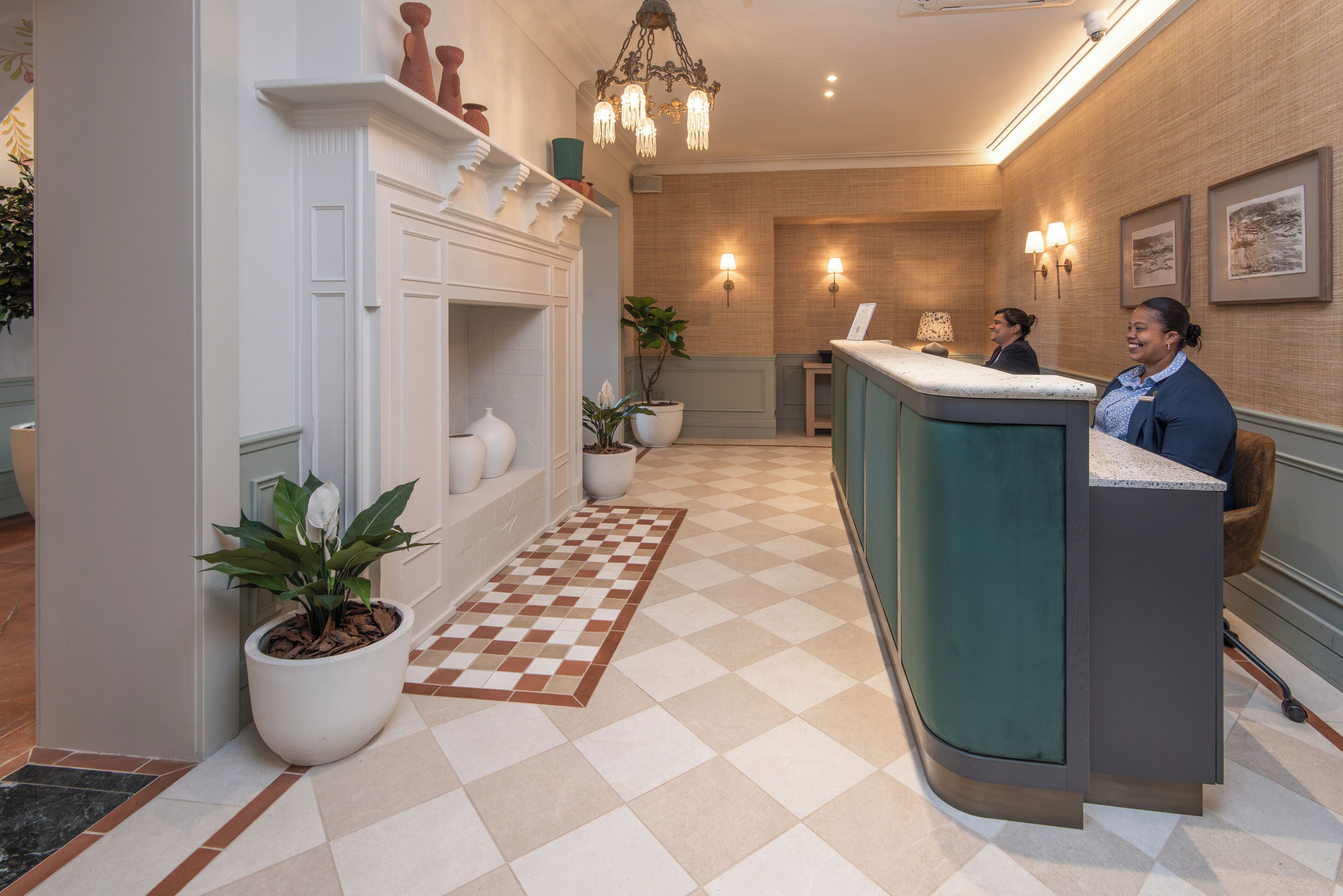 Un espace d’accueil avec un sol à carreaux, un comptoir de réception, des plantes en pot, une cheminée et des lampes fixées au mur.