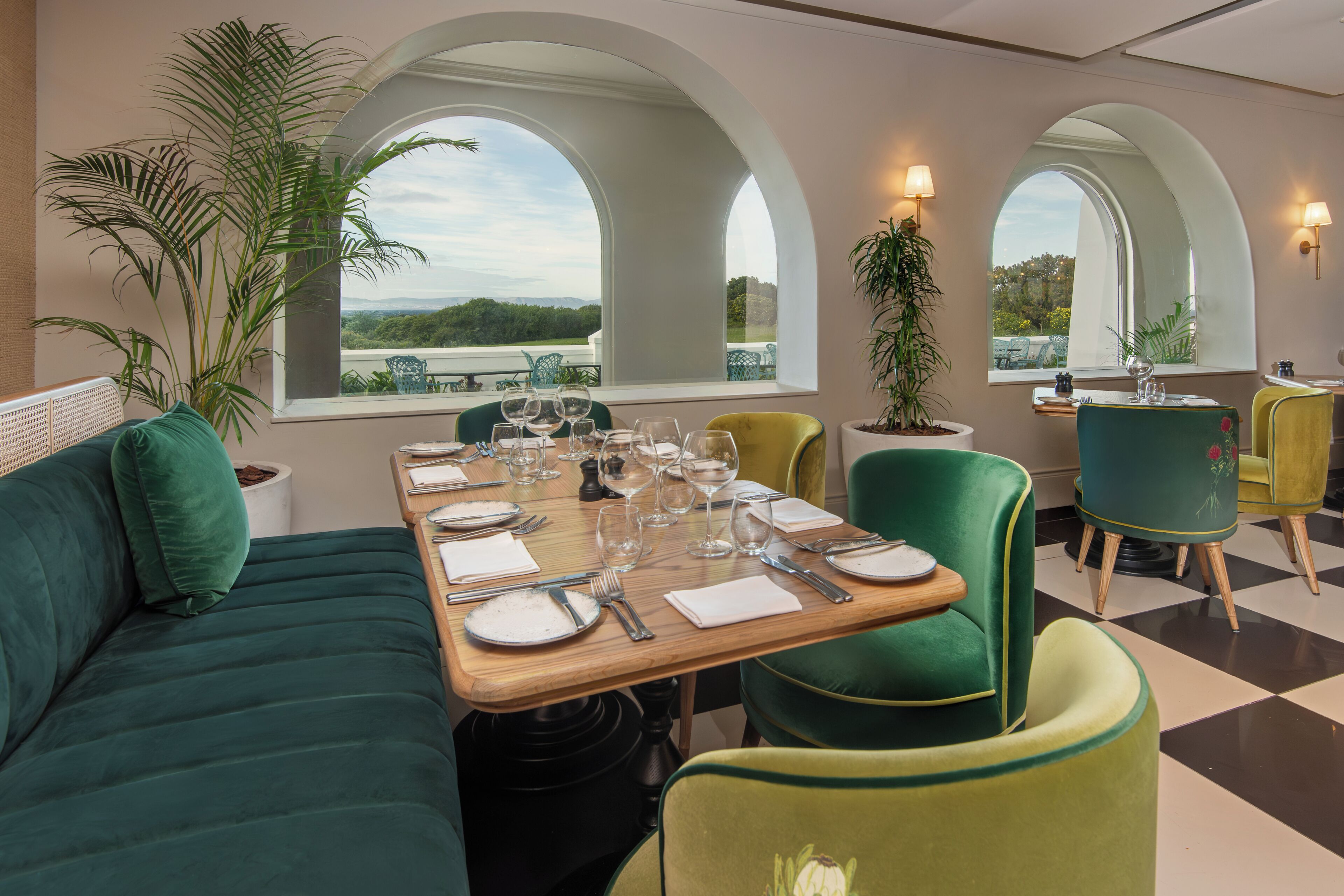 Un comedor con una mesa dispuesta para cuatro personas, sillas tapizadas de verde y amplias ventanas en arco que dan a vistas de follaje.