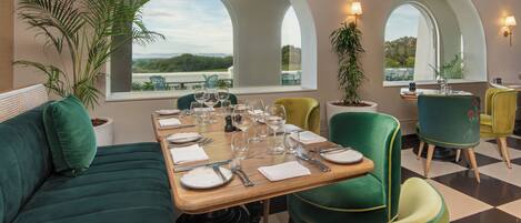 Un comedor con una mesa dispuesta para cuatro personas, sillas tapizadas de verde y amplias ventanas en arco que dan a vistas de follaje.