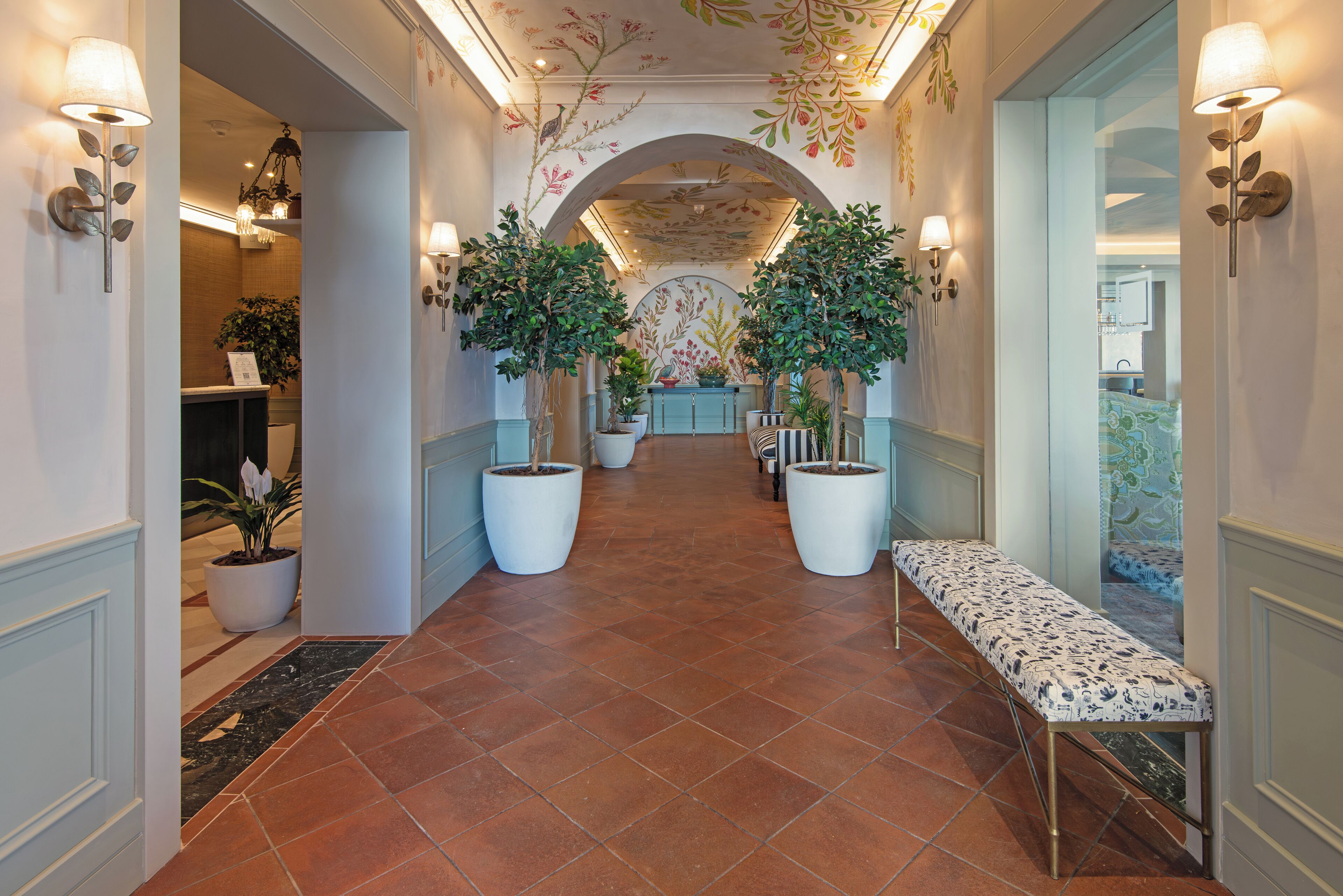 A hallway with large potted plants, a bench, and wall-mounted lighting fixtures.
