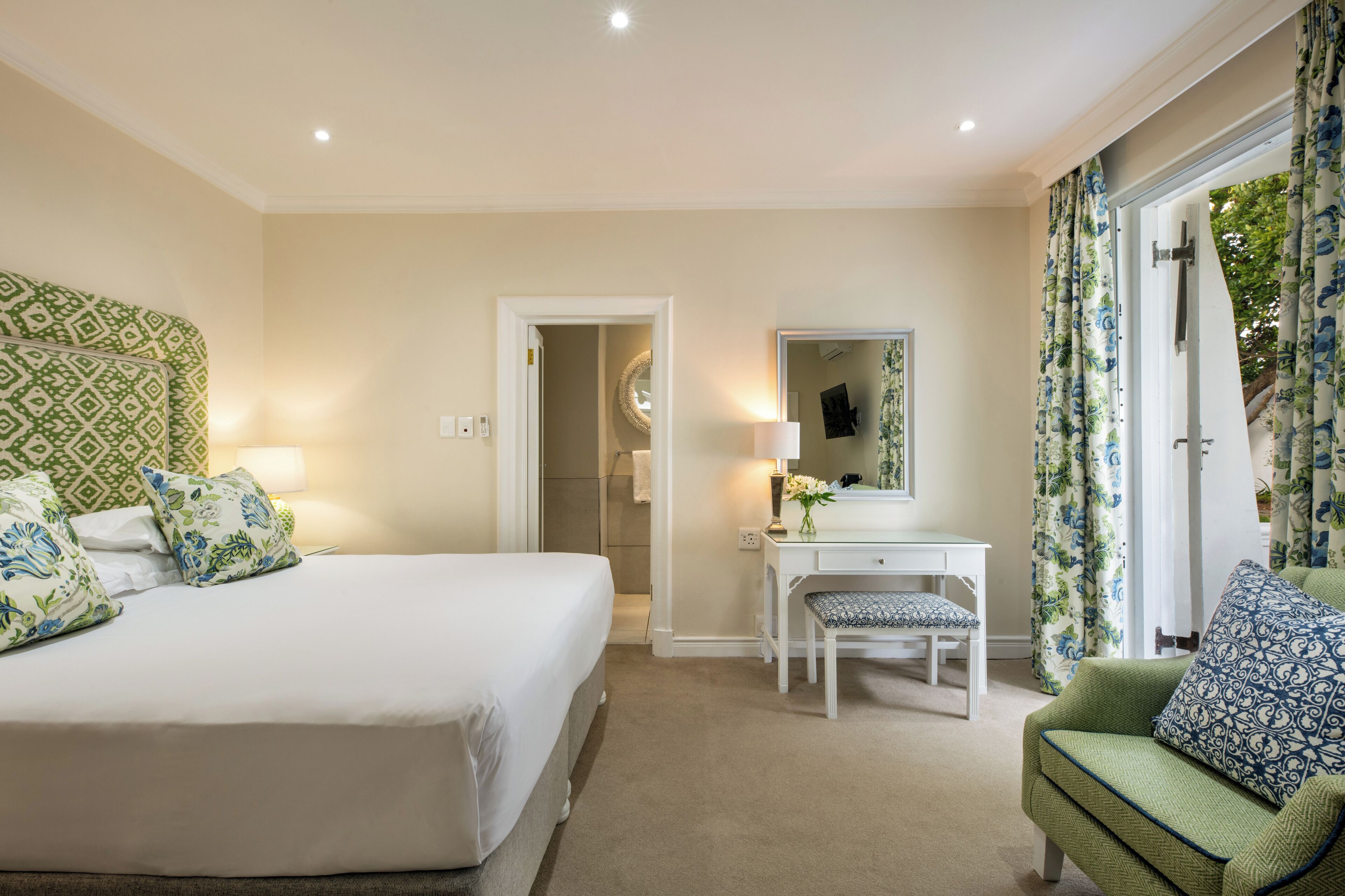 A hotel room with a large bed, a small desk, a chair, and a sofa. There is a mirror above the desk and a door leading to another room.