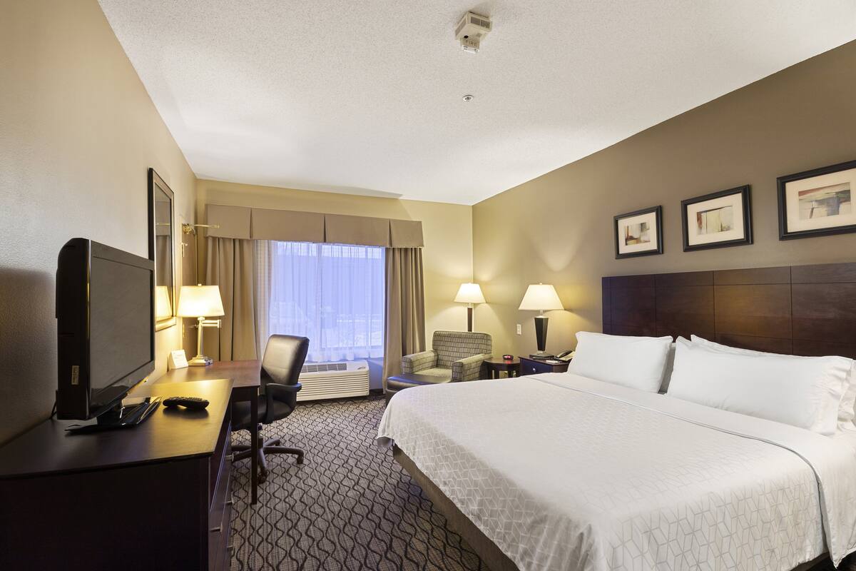 A hotel room with a large bed, a desk with a computer and phone, a chair, a TV, and two lamps.