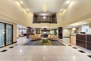 Un'ampia hall con soffitto alto, un'area salotto centrale con divani, una reception e una scala che conduce a un piano superiore.