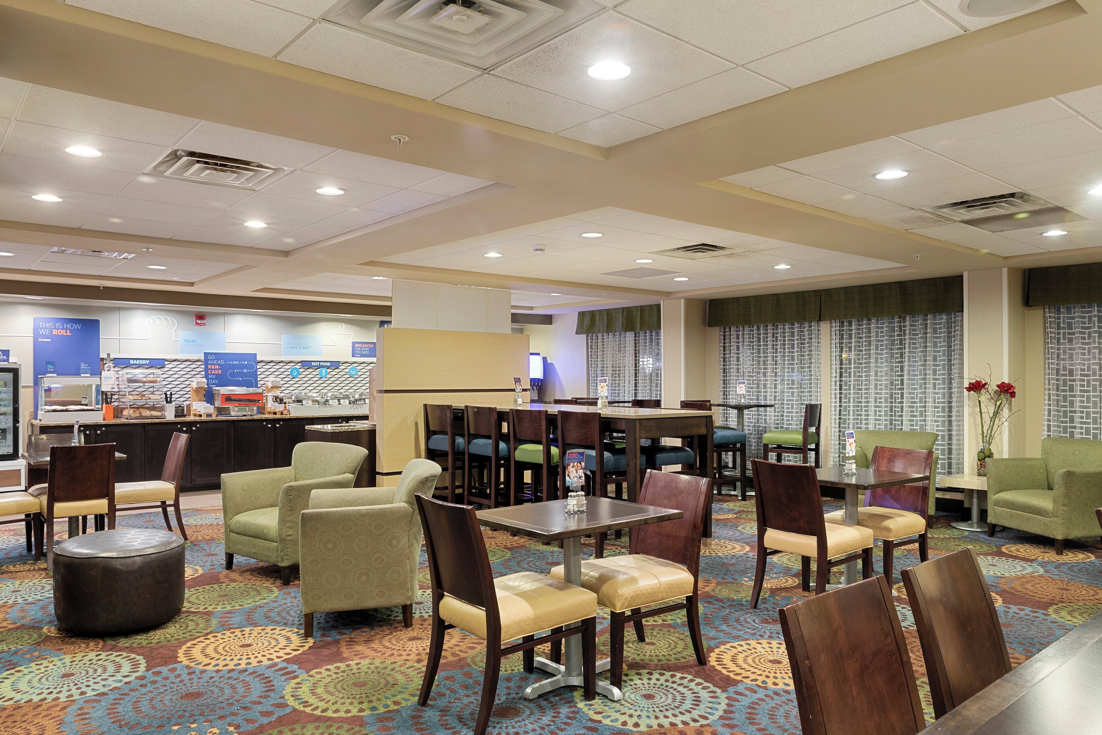 A hotel dining area with multiple tables and chairs, a bakery counter, and a view of the cityscape through large windows.
