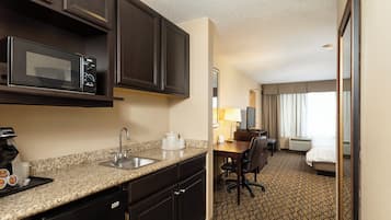 A hotel room with a kitchenette, including a microwave, sink, and coffee maker. There is a desk with a chair, a bed, and a window with curtains.