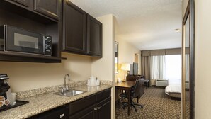 A hotel room with a kitchenette, including a microwave, sink, and coffee maker. There is a desk with a chair, a bed, and a window with curtains. - Holiday Inn Express Bloomington West by IHG (Bloomington)