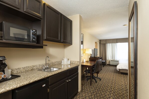 A hotel room with a kitchenette, including a microwave, sink, and coffee maker. There is a desk with a chair, a bed, and a window with curtains. - Holiday Inn Express Bloomington West by IHG (Bloomington)