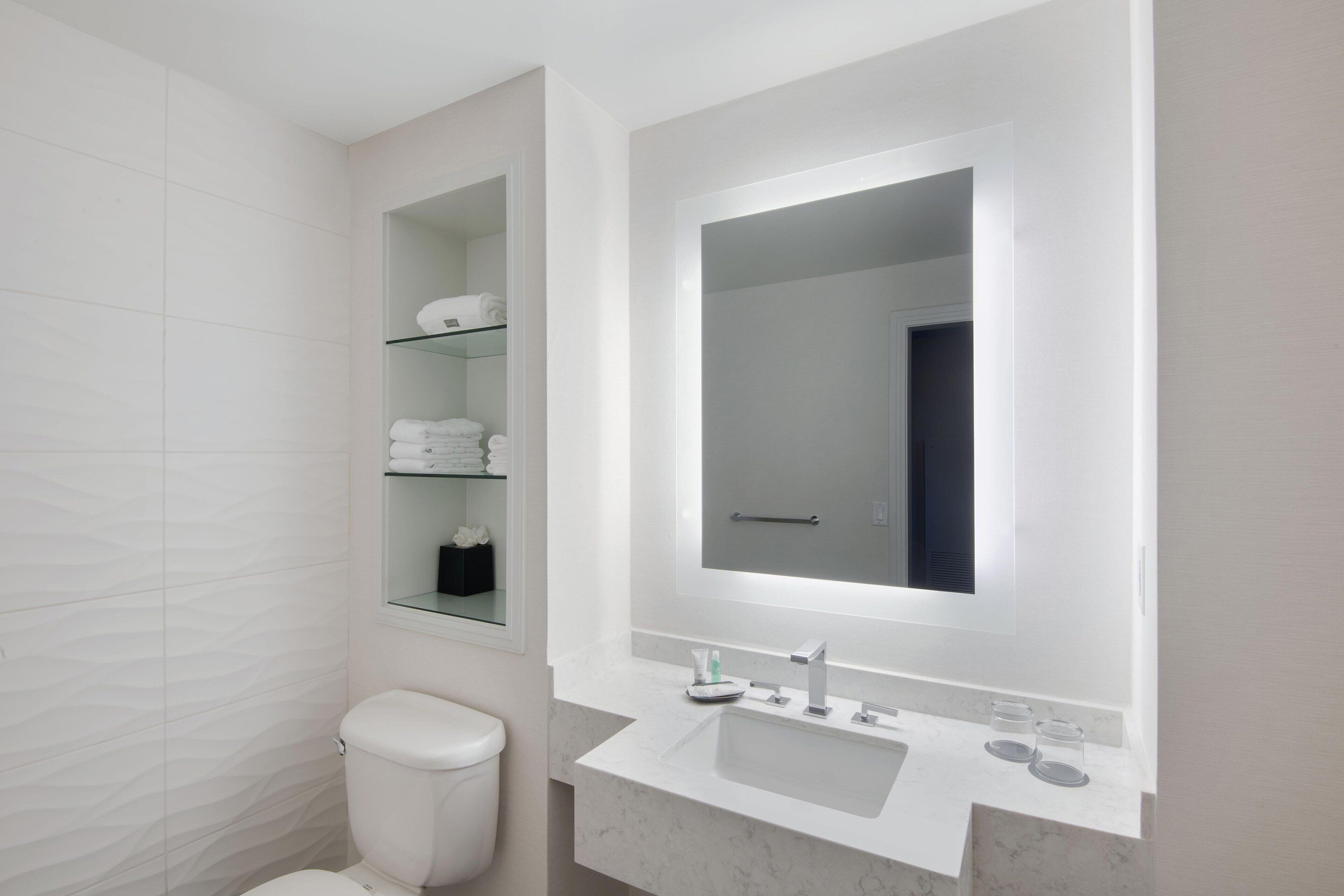 A modern bathroom with a toilet, a sink, a mirror, and a glass shelf with towels.