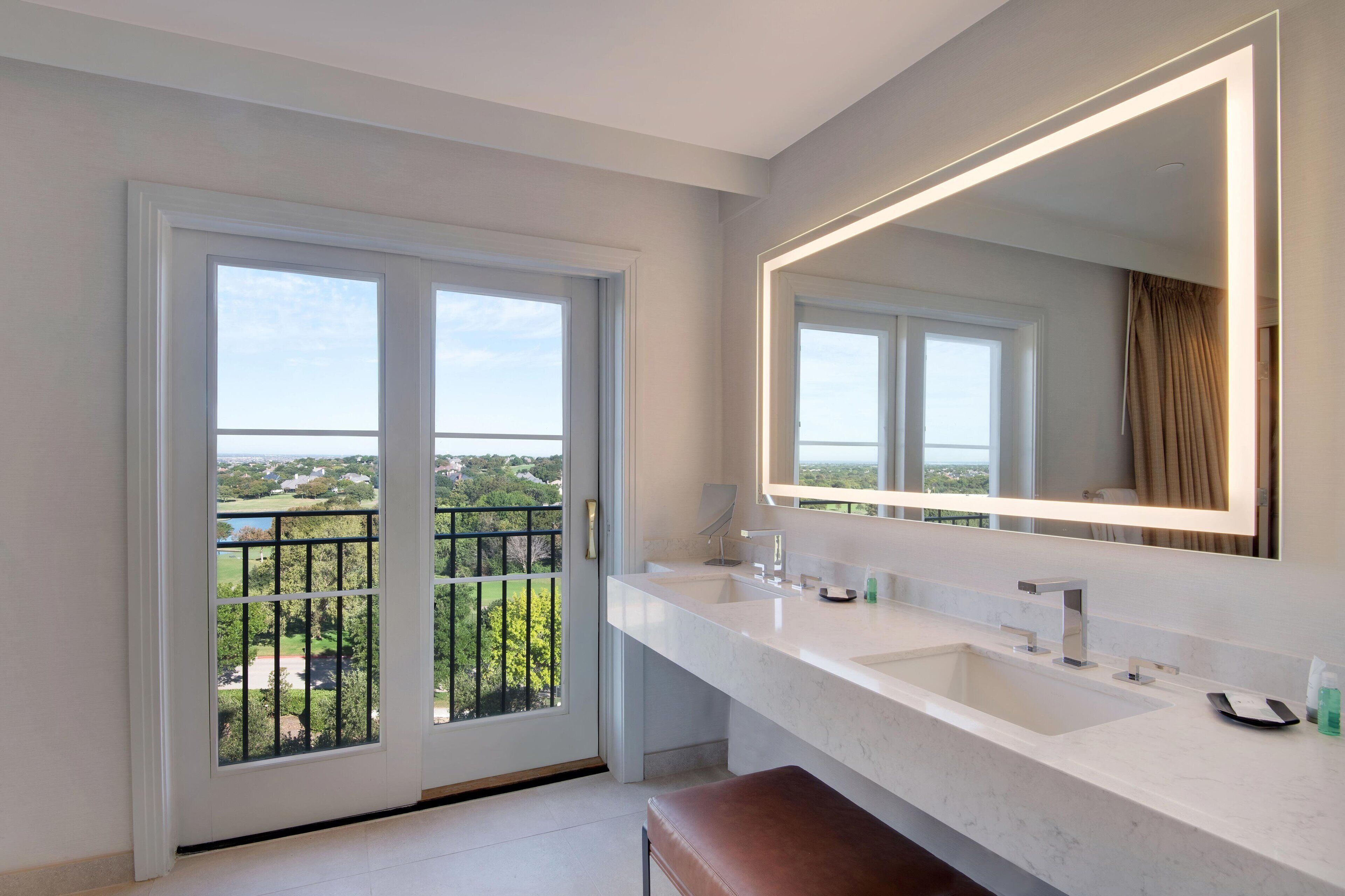 A modern bathroom with a large mirror, two sinks, and a view of greenery through the door.