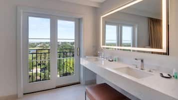 A modern bathroom with a large mirror, two sinks, and a view of greenery through the door.