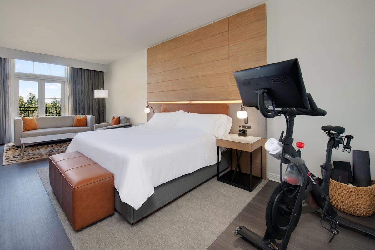 A modern hotel room with a bed, a desk with a computer, a standing exercise bike, and a view of the outdoors.