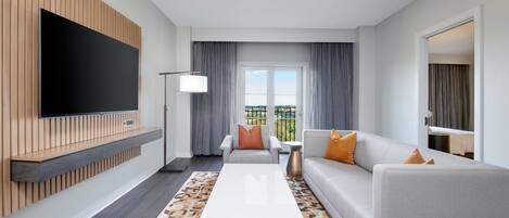 A modern living room with a flat-screen TV mounted on a wooden panel, a grey sofa with orange cushions, a white coffee table, and a patterned rug.