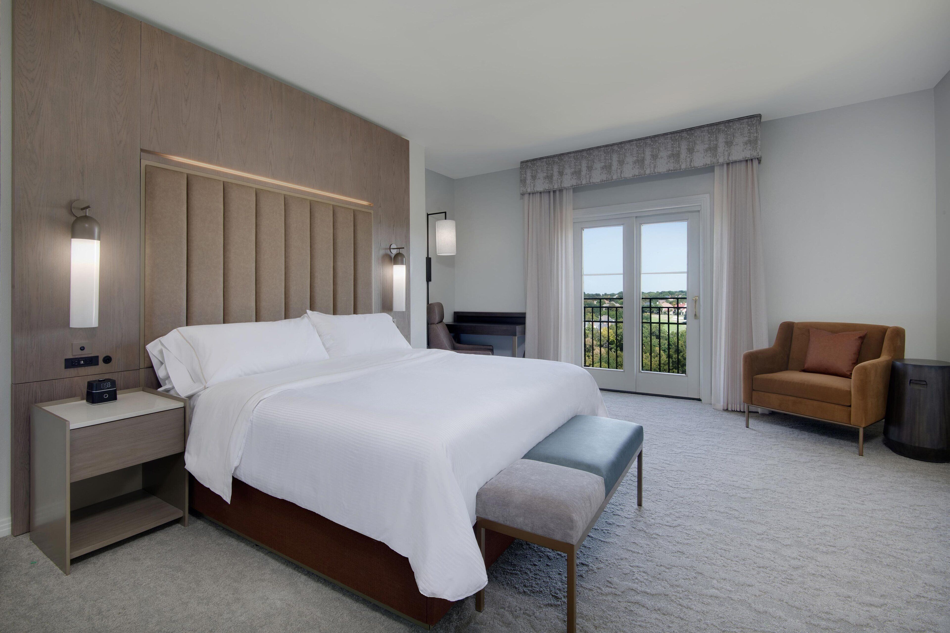 Un hotel moderno con una cama grande, mesitas de noche, una silla y vistas al exterior.