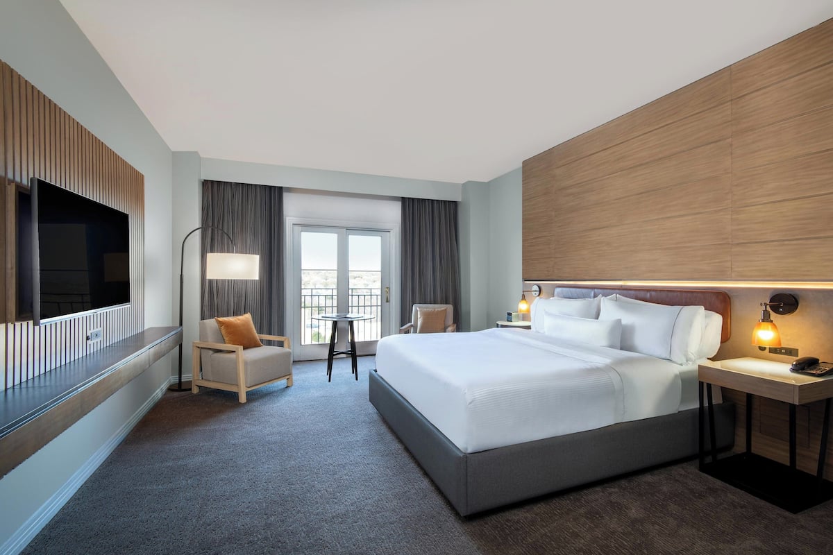 A hotel room with a large bed, a TV mounted on the wall, a desk with a lamp, and a view of the outdoors through a window.