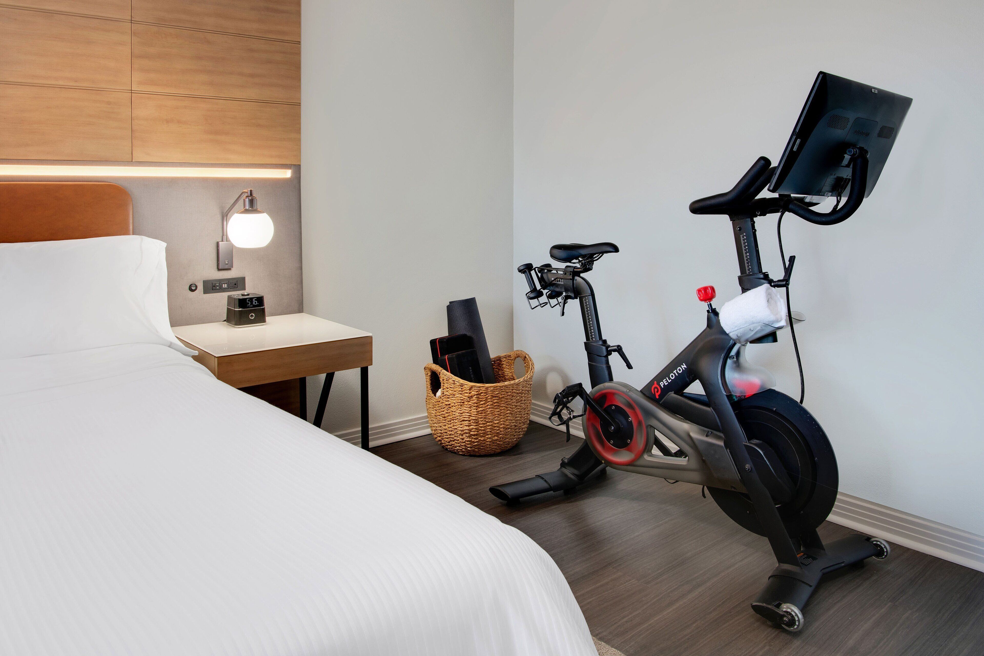 Et hotelværelse med en seng, et skrivebord, en vægmonteret lampe, en Peloton træningscykel og en fletkurv.