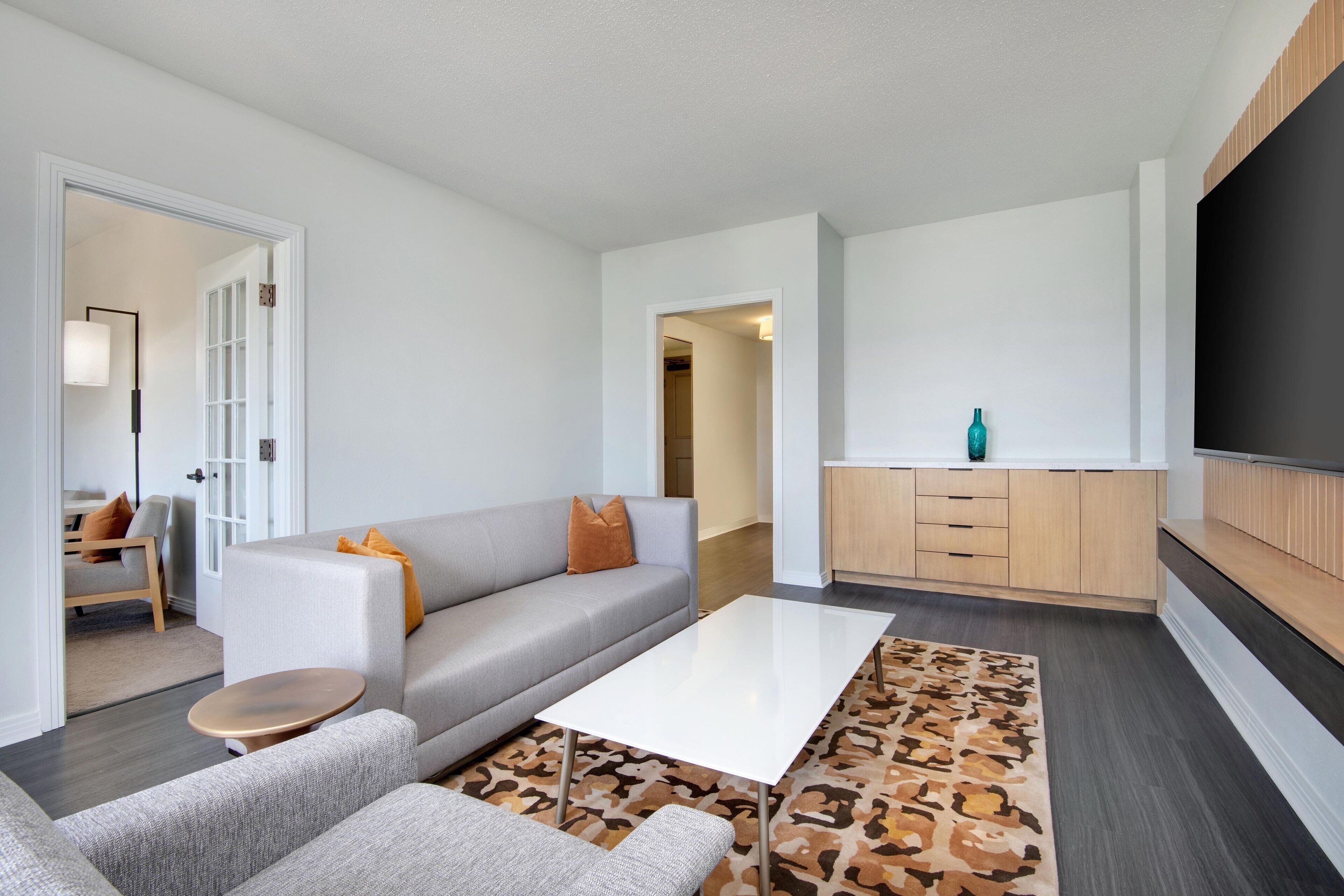 A modern living room with a grey sofa, a coffee table, and a patterned rug. There is a kitchen area with wooden cabinets and a teal bottle on the counter.