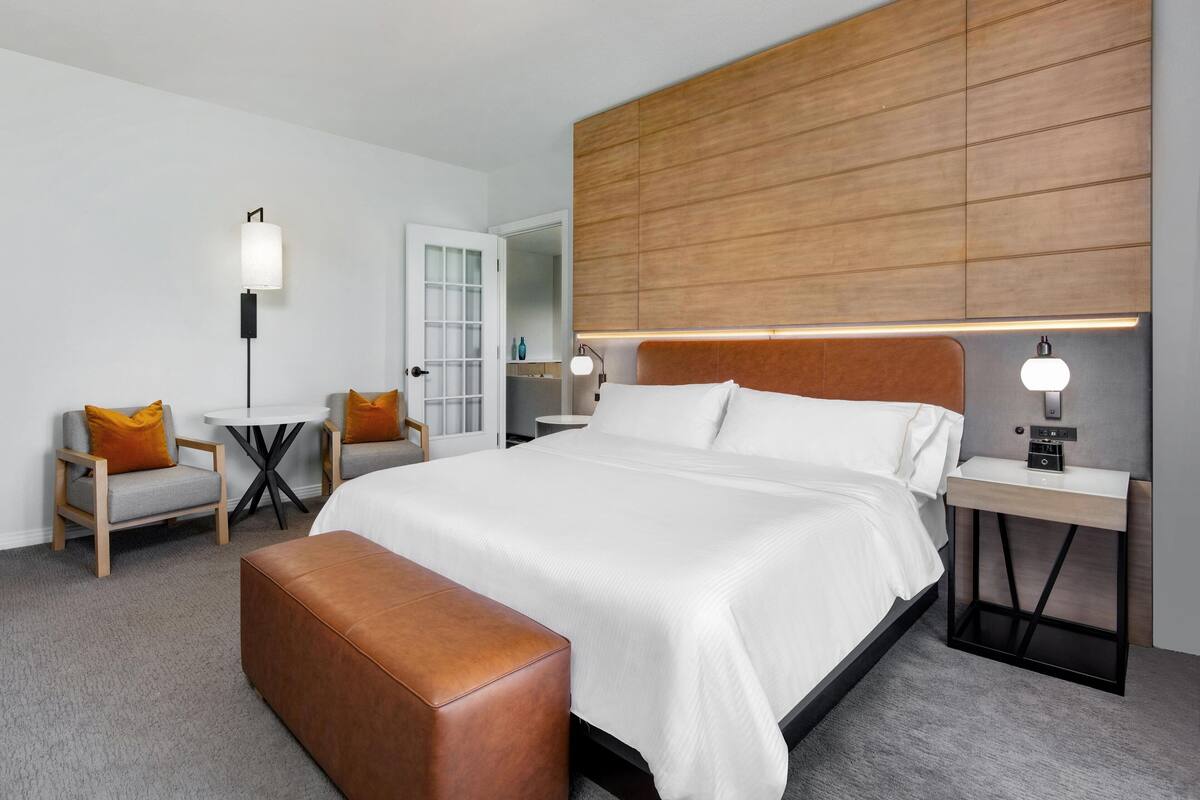 A modern hotel room with a large bed, a wooden headboard, a small table, a bench, and a nightstand with a lamp.