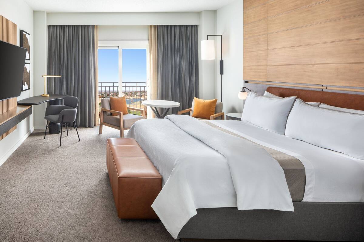 A hotel room with a large bed, a desk, a chair, a small table, and a balcony with a view.