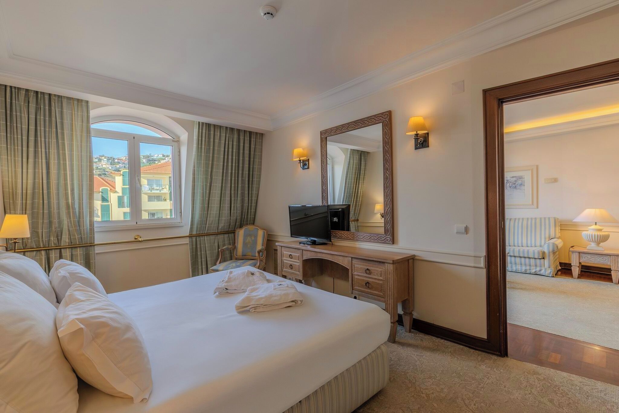A hotel room with a bed, a desk with a television, a mirror, and a view of the outside.