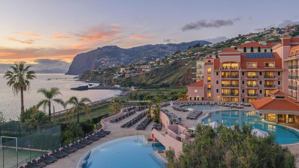 Un resort con una grande piscina, lettini prendisole e vista sull'oceano e sulle colline.
