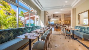 Un ristorante spazioso con soffitti alti, ampie finestre e vista su palme. I tavoli sono apparecchiati con sedie e tavoli da pranzo.