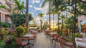Zona pranzo all'aperto con tavoli e sedie, circondata da piante tropicali e palme.
