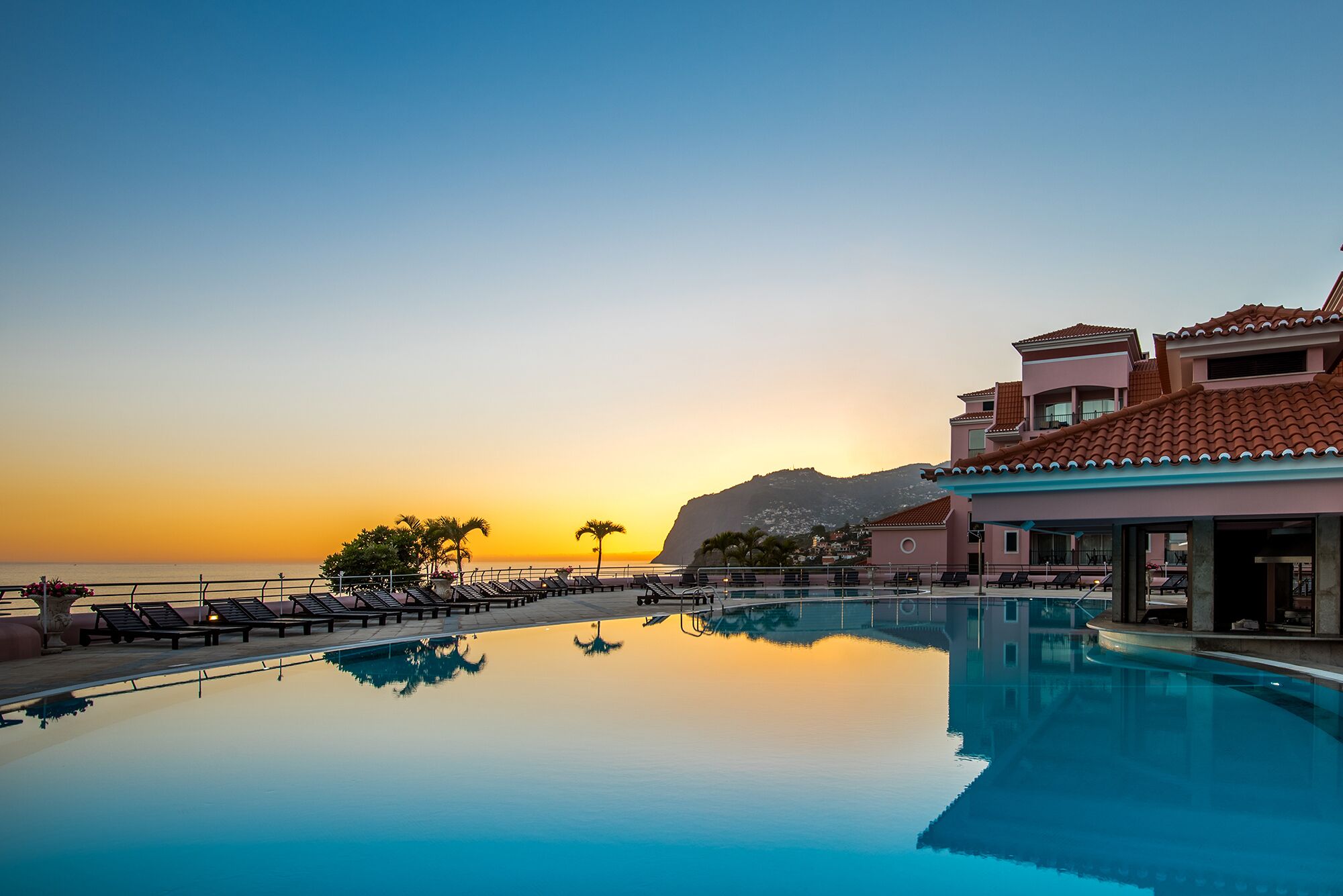 Une piscine d’hôtel avec des chaises longues, un ciel dégagé et une montagne en arrière-plan.