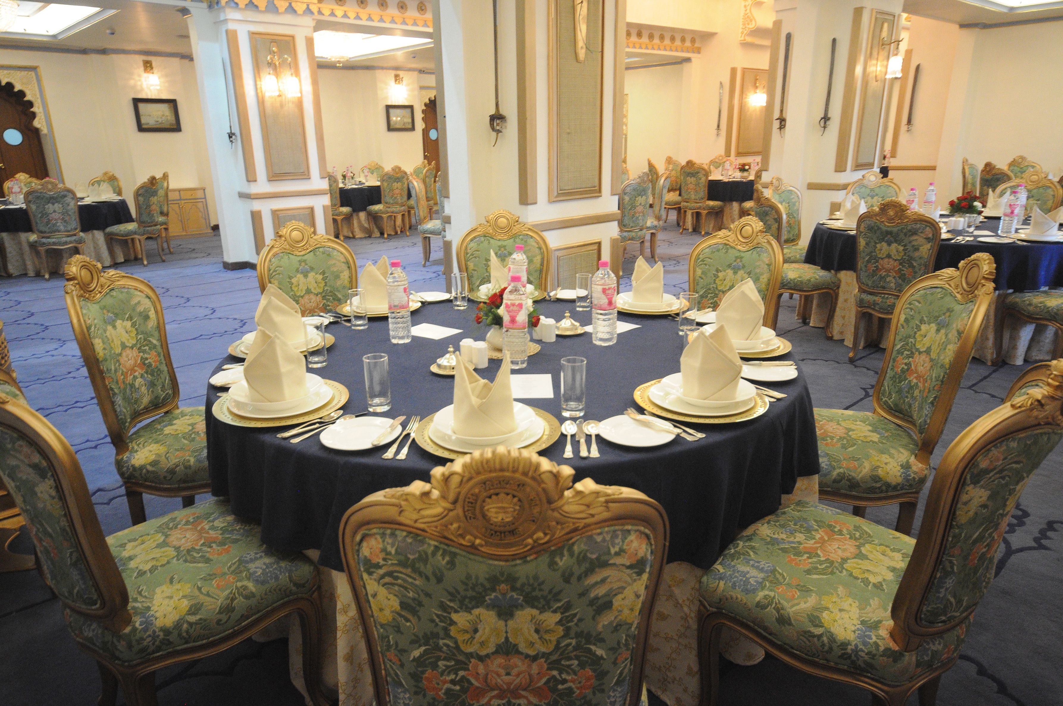 Une salle de banquet avec des tables rondes dressées pour le repas, chacune agrémentée de chaises décorées et recouverte d’une nappe sombre.