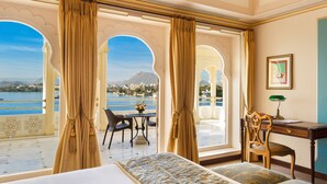 A hotel room with a large bed, a desk, and a view of a body of water and mountains. - Taj Fateh Prakash Palace (Udaipur)