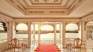 A room with a red carpet, wooden chairs, and a view of a body of water and buildings. - Taj Fateh Prakash Palace (Udaipur)