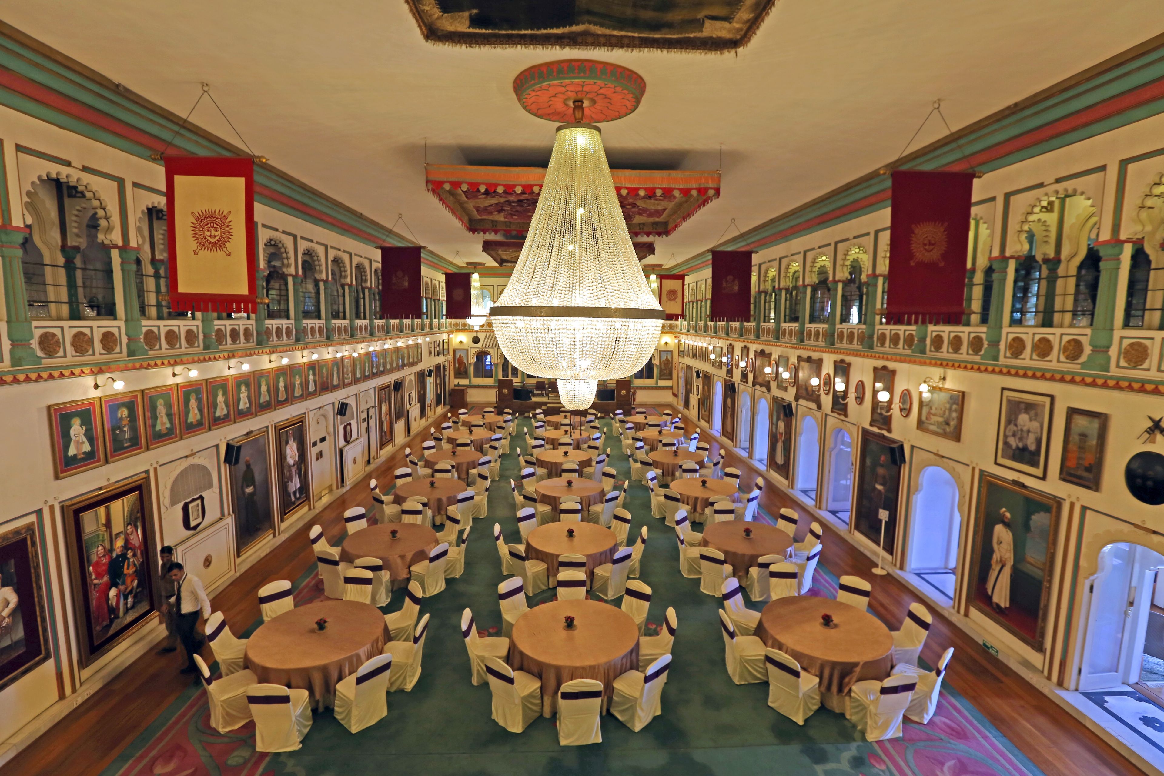 A grand hall with multiple round tables and chairs, ornate ceiling, and framed artwork on the walls.