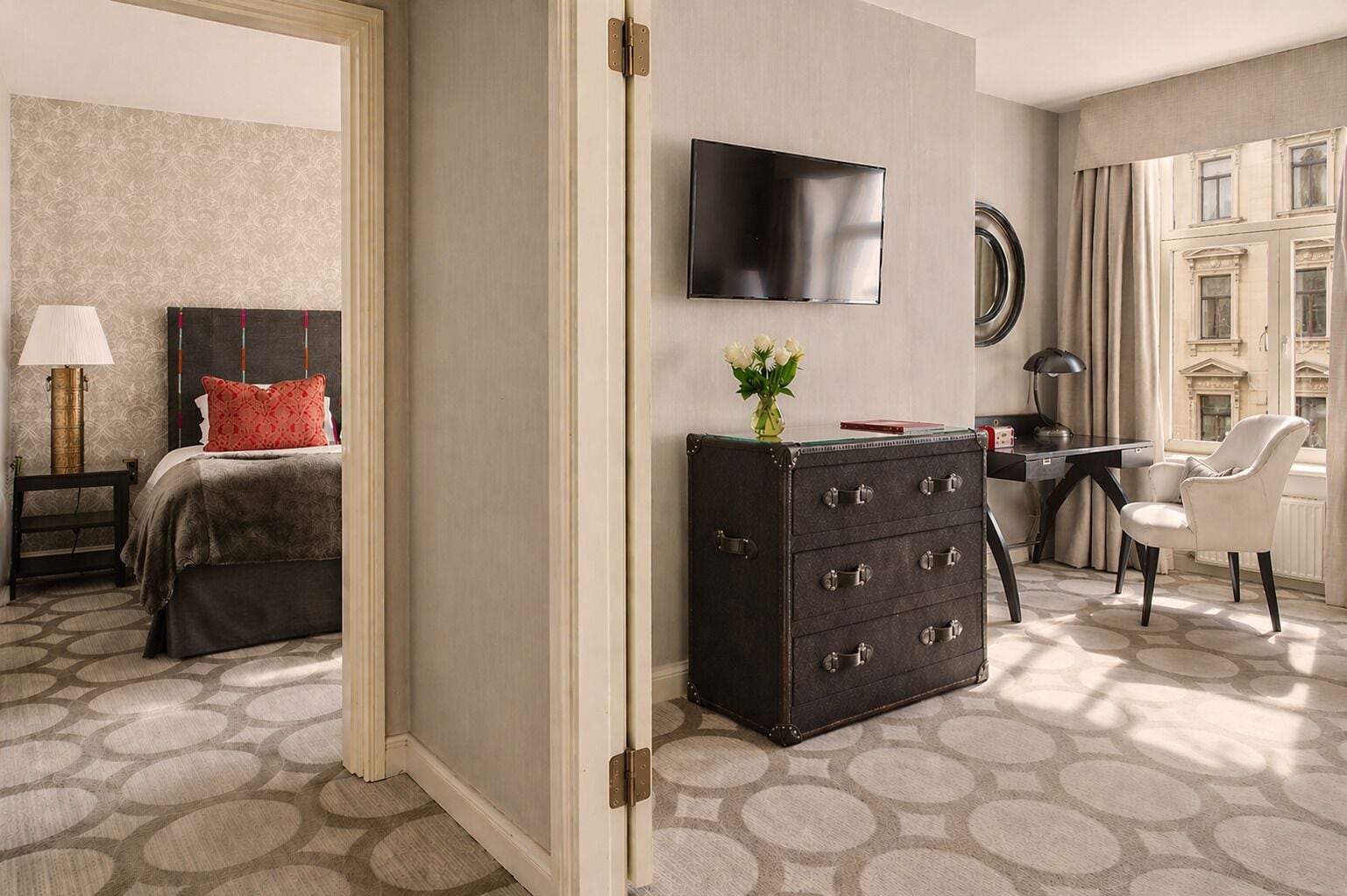 A hotel room with a bed, a desk, a dresser, a TV, and a view of a building through the window.