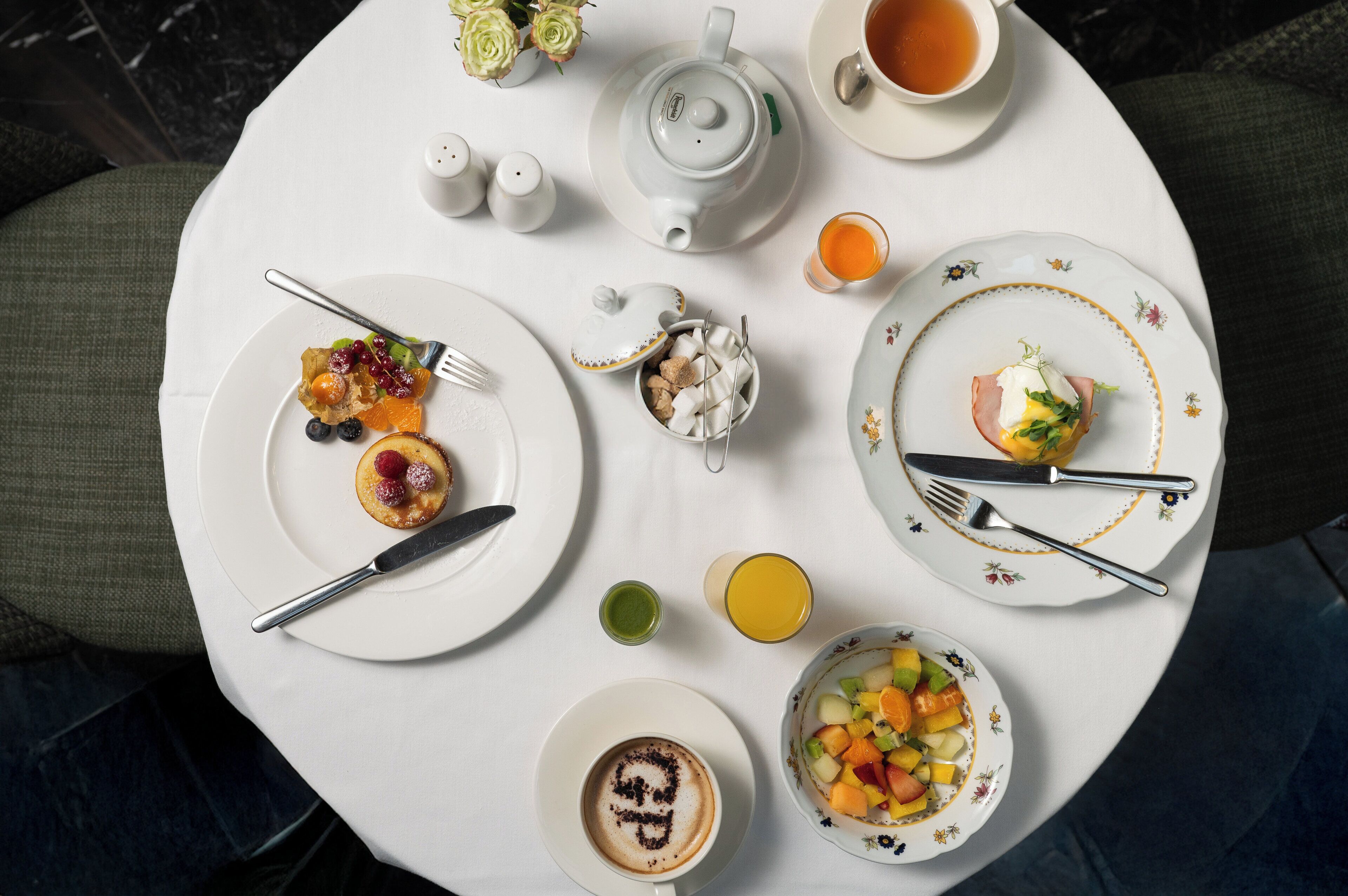 A round dining table with various breakfast items including a teapot, cups, plates with food, and utensils.
