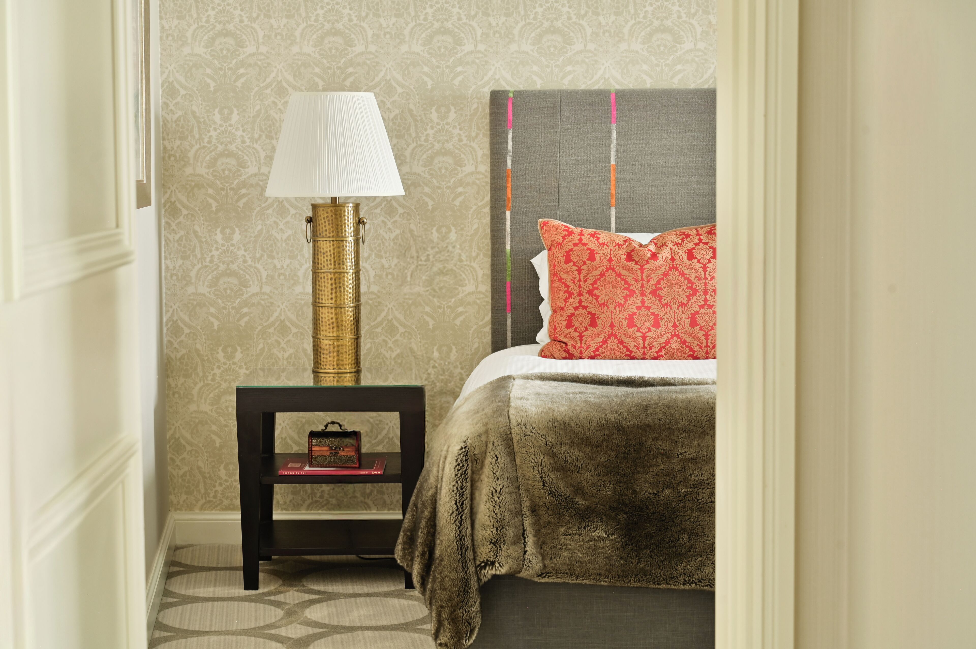 A neatly arranged bedroom with a bed, a bedside table with a lamp, and a small shelf with books.