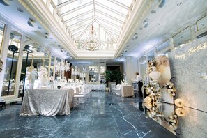 A spacious, well-lit hall with a high ceiling and a central chandelier, tables set for a formal event, and a large mirror on one wall.